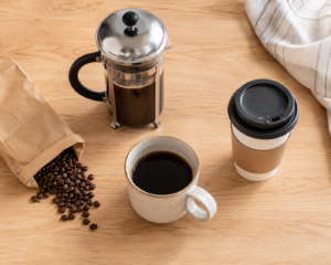 A close-up, angled shot of freshly ground coffee beans in a grinder next to a finished cup of coffee, on a light wooden table with soft natural morning light. No text, no logos. Style: warm, clean, premium editorial lifestyle photography, realistic.